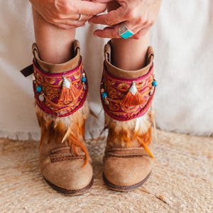 Könnte beinhalten: Braune Wildleder-Stiefeletten mit einer dekorativen Manschette mit dunkelrotem und goldenem Paisley-Muster, orangefarbenen Quasten, kleinen silbernen Anhängern und türkisfarbenen Perlen. Die Stiefel sind mit braunen und weißen Federn verziert.