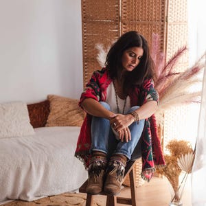 May include: A woman wearing a red, black and white patterned cardigan with blue jeans and brown fringed boots sits on a wooden stool. She is looking down and has long dark hair.