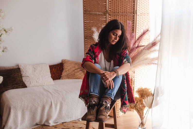 May include: A woman wearing a red, black and white patterned cardigan with blue jeans and brown fringed boots sits on a wooden stool. She is looking down and has long dark hair.