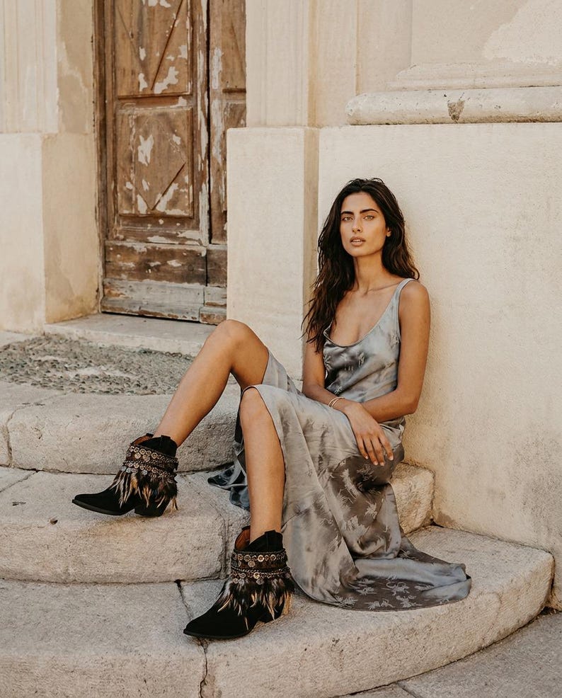 May include: A sleeveless grey tie-dye maxi dress with a scoop neckline paired with black ankle boots featuring decorative metallic coin embellishments and brown feather fringe accents around the top of the boot shaft.