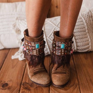 May include: Brown suede ankle boots with colorful patterned fabric and turquoise gemstone accents. The boots have a fringe detail and are worn on a wooden floor.