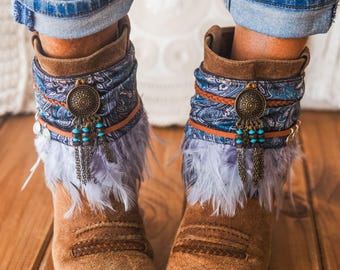 Op de afbeelding: Bruine suède enkellaarsjes met een blauwe manchet van paisley-stof, lichtpaarse veren en een centrale bronskleurige ronde bedel met turquoise kralen en metalen kettinkjes. De laarsjes worden gedragen met een blauwe spijkerbroek met omgeslagen pijpen.