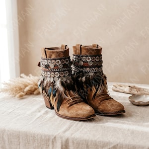 May include: A pair of tan suede ankle boots featuring a decorative cuff adorned with metallic coin charms, a thin silver-toned chain, and a fringe of brown and black feathers. The boots have a rounded toe, a stacked wooden heel, and a textured finish.