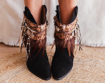May include: A pair of black ankle boots featuring a pointed toe and a decorative cuff. The cuff is adorned with a brown patterned fabric, brown feathers, silver-toned metal charms, beads, and thin leather-like fringe strips hanging down.