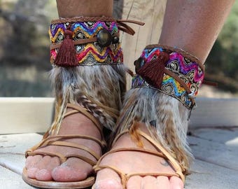 Op de afbeelding: Een paar beige leren sandalen met enkelbanden versierd met veelkleurig zigzagborduurwerk, bruine kwastjes en donkere ronde accenten. De enkelbanden zijn voorzien van lange, natuurlijke veren die over de voeten vallen.