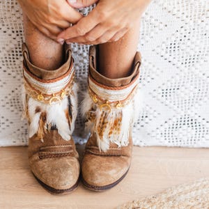 May include: A pair of tan suede ankle boots featuring decorative cuffs adorned with white and brown feathers, braided fabric trim, and small gold-toned circular metal charms hanging from the top edge of the boot shafts.