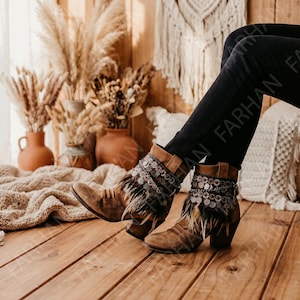 May include: A pair of brown suede ankle boots featuring a stacked heel, a rounded toe, and decorative fringe detailing around the ankle. The boots are adorned with a band of small, metallic circular coins and dark feathers layered over the suede.