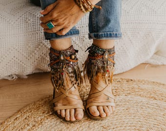 May include: A pair of tan leather sandals featuring ankle cuffs decorated with patterned fabric, brown braided cords, metallic charms, and dangling brown and black feathers. The sandals have multiple thin straps across the foot and open toes.
