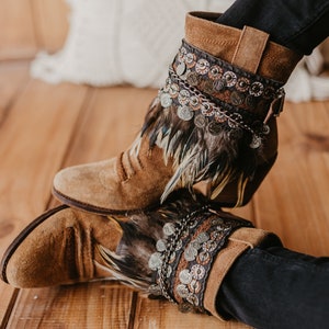 May include: Brown suede ankle boots featuring a decorative cuff adorned with layered dark feathers, metallic coin charms, and intricate braided trim. The boots have a rounded toe, a stacked heel, and a side buckle detail on the upper shaft.