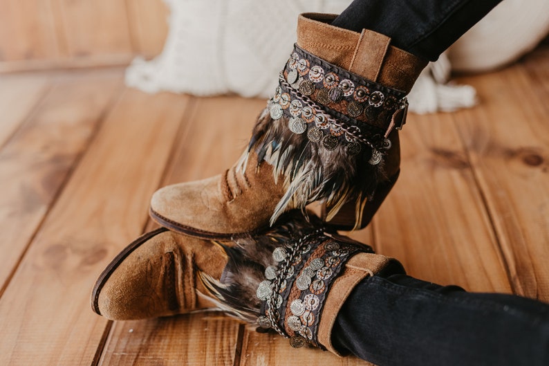May include: Brown suede ankle boots featuring a decorative cuff adorned with layered dark feathers, metallic coin charms, and intricate braided trim. The boots have a rounded toe, a stacked heel, and a side buckle detail on the upper shaft.