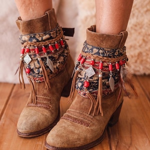 May include: A pair of brown suede ankle boots featuring decorative wraps around the cuffs. The wraps include dark fabric with a floral pattern, braided leather cords, bright red cylindrical beads, silver-toned charms, and brown fringe tassels.