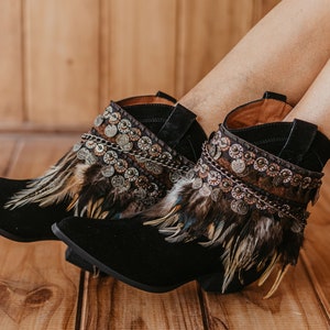 May include: Black ankle boots featuring a pointed toe and a low block heel. The upper shaft is adorned with a decorative band of metallic circular coins, dark chains, and layered brown and black feathers that wrap around the ankle area.