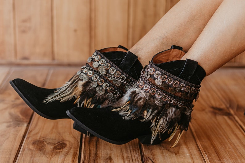 May include: Black ankle boots featuring a pointed toe and a low block heel. The upper shaft is adorned with a decorative band of metallic circular coins, dark chains, and layered brown and black feathers that wrap around the ankle area.