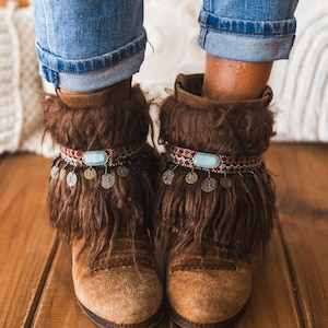 May include: A pair of brown suede ankle boots featuring long brown faux fur cuffs. Each boot is adorned with a decorative band featuring a central light blue oval stone and a series of small, dangling metallic coin charms around the ankle.