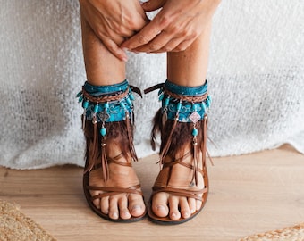 Op de afbeelding: Bruine leren sandalen met enkelmanchetten van blauwgroene stof met patroon, versierd met turkooiskleurige kralen, zilverkleurige bedels en bruine franjes. De sandalen hebben dunne bandjes die over de bovenkant van de voet lopen.