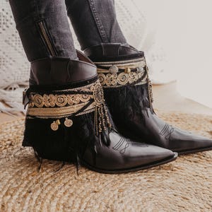 May include: Black leather ankle boots featuring a pointed toe and a decorative cuff. The cuff is adorned with intricate gold-tone embroidery, dangling metallic coin charms, and black feathers. The boots have a side buckle and stitched detailing.