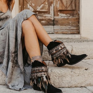 May include: Black ankle boots featuring a pointed toe, a low block heel, and decorative embellishments around the collar. The boots are adorned with a band of metallic coins and layered brown and black feathers hanging from the top edge.