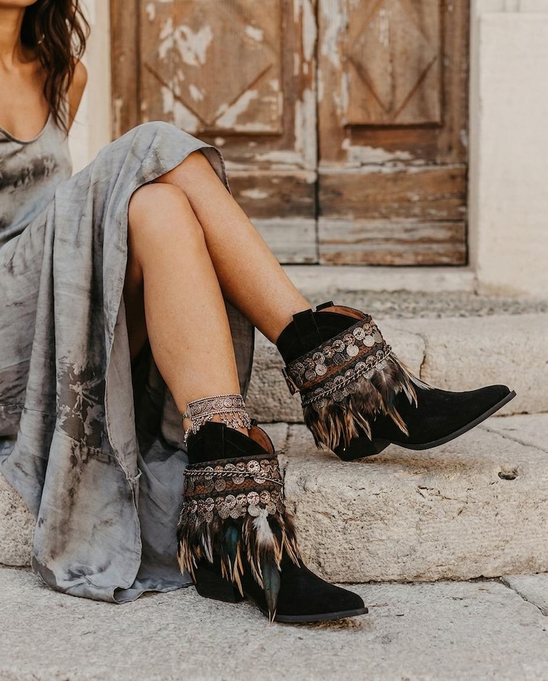 May include: Black ankle boots featuring a pointed toe, a low block heel, and decorative embellishments around the collar. The boots are adorned with a band of metallic coins and layered brown and black feathers hanging from the top edge.