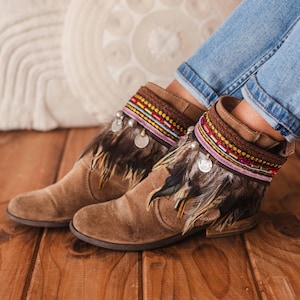 Puede incluir: Botas de tobillo de ante marrón con ribete de plumas y adornos de cuentas de colores. Las botas las lleva una persona que lleva pantalones vaqueros azules.