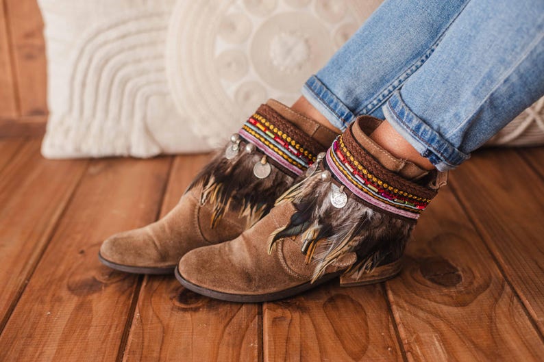 May include: Brown suede ankle boots with feather trim and colourful beaded embellishments. The boots are worn by a person wearing blue jeans.