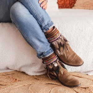 May include: Brown suede ankle boots with colourful beaded and feathered embellishments. The boots are being worn by a person sitting on a white fluffy surface.