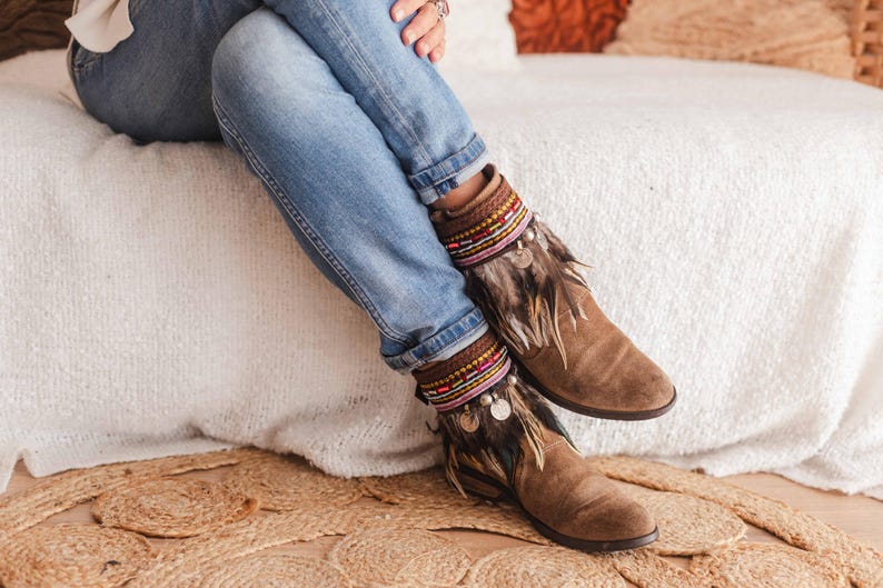 May include: Brown suede ankle boots with colourful beaded and feathered embellishments. The boots are being worn by a person sitting on a white fluffy surface.