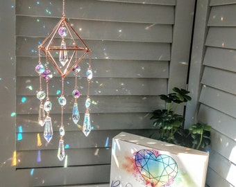 May include: A copper wire geometric suncatcher with iridescent crystals hanging from it. The suncatcher is hanging from a white ceiling. The suncatcher is casting rainbows on a white wall and a box with the text "Rainbows and Wishes Suncatchers to Brighten Your Life".