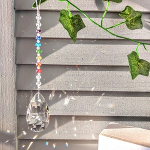 May include: A rainbow crystal suncatcher hanging from a white string. The suncatcher is made of clear glass beads and has a teardrop-shaped crystal at the bottom. The suncatcher is hanging in front of a white wooden wall with green ivy vines.