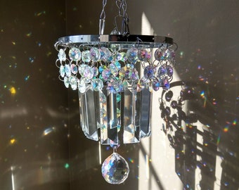 May include: A silver crystal chandelier with a round base and a teardrop crystal hanging from the bottom. The chandelier is hanging from a chain and casts rainbows on the wall.