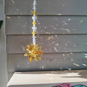 May include: A crystal suncatcher with a yellow and clear crystal starburst hanging from a white string. The suncatcher is hanging in front of a white wooden slat wall.