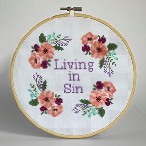 Puede incluir: Arte de bordado en aro con fondo blanco y texto morado que dice "Living in Sin". El aro está decorado con flores rosas y moradas.