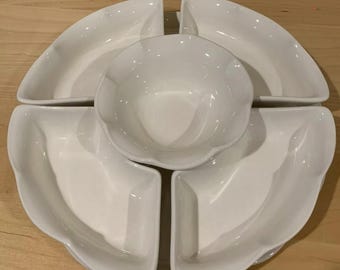 May include: A white ceramic serving dish set. The set includes a central bowl surrounded by four curved, petal-shaped dishes. The pieces have a glossy finish and are arranged on a light-colored wooden surface.