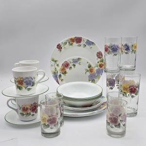 May include: A collection of white dinnerware with a floral pansy design. The set includes plates, bowls, cups, saucers, and drinking glasses. The pansies are in shades of purple, yellow, and red.