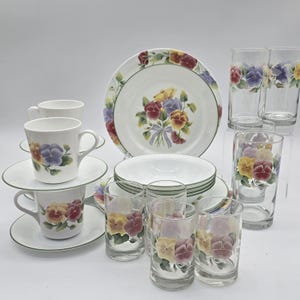May include: A complete floral dinnerware set featuring white plates, bowls, cups, saucers, and drinking glasses. Each piece is decorated with a colorful pansy design in shades of yellow, purple, and red, with green trim.