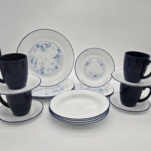 May include: A white dinnerware set with blue floral accents and navy blue trim. The set includes plates, saucers, bowls, and mugs. The plates and bowls have a blue border. The mugs are navy blue.