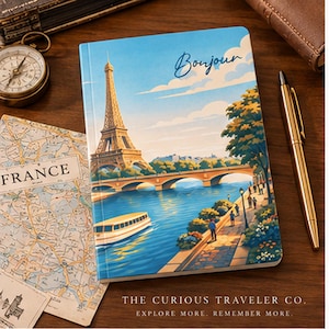 May include: A rectangular notebook featuring a colorful illustration of the Eiffel Tower, a river, a boat, and people walking along a path, with the word Bonjour printed at the top. The notebook is surrounded by a map of France, a compass, and a gold pen.