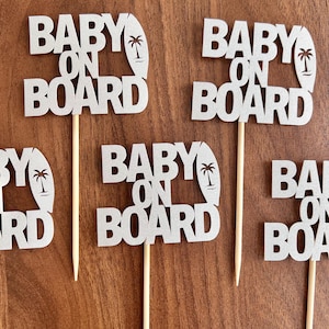 May include: Several glittery silver "Baby on Board" cake toppers. Each topper features the text "Baby on Board" with a surfboard and palm tree design. The toppers are attached to wooden sticks for easy insertion into cakes or other treats.