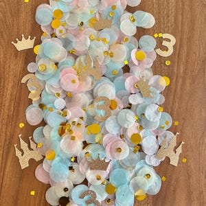 May include: A collection of pastel-colored confetti, including pink, blue, and yellow circles, scattered on a wooden surface. Gold glitter crowns, castles, and the number "3" are interspersed throughout the confetti, creating a festive and celebratory atmosphere.