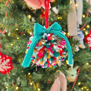 May include: A colorful yarn pom-pom ornament with a teal ribbon bow. The ornament is hanging from a red string.