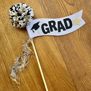 May include: A white and black pom-pom with gold glitter accents attached to a wooden stick. A white banner with black text that says "GRAD" and a gold glitter star is attached to the stick.