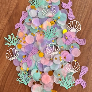 May include: A collection of pastel-colored confetti, including pink, purple, and teal circles, with gold accents. Decorative cutouts of mermaid tails, seashells, and coral are scattered throughout. The confetti is arranged on a wooden surface.