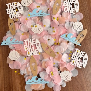 May include: A collection of confetti in shades of pink, white, and peach, with decorative elements. The design includes cutouts of surfboards, seashells, and waves, along with the text "THE BIG ONE".