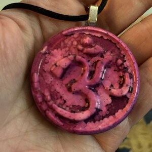 May include: A circular pendant with a pink and purple marbled pattern, featuring the Om symbol. It hangs from a black cord necklace with a silver-coloured clasp.