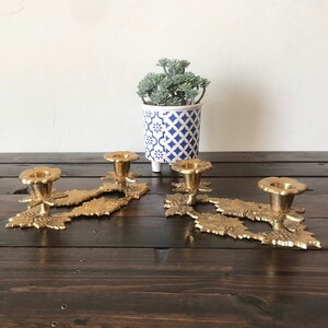May include: A set of three gold-toned brass candle holders with a leaf design. The candle holders are on a dark wood surface.