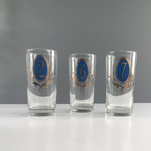 May include: Three clear drinking glasses with blue oval designs featuring the numbers 2, 6, and 7. The numbers are framed with gold filigree. The glasses are tall and cylindrical.