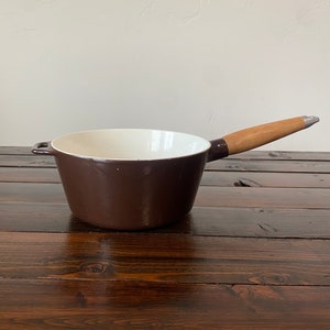 May include: A brown enameled cast iron saucepan with a wooden handle. The saucepan has a white interior and is sitting on a wooden surface.