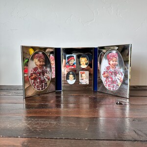 May include: A silver-toned, tri-fold picture frame with oval and rectangular openings. The frame holds several small photographs, including two oval portraits. The frame is displayed on a dark wooden surface.