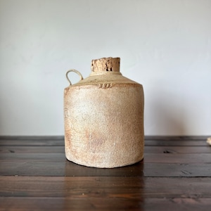May include: A beige ceramic jug with a cork stopper and a handle. The jug has a rustic, handmade look and is sitting on a dark wooden surface.