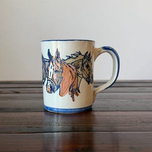 May include: A cream-colored ceramic mug featuring a hand-painted illustration of three horses in shades of brown, tan, and black. The mug has a blue rim, a blue base border, and a blue-accented handle, showcasing a glossy finish throughout.