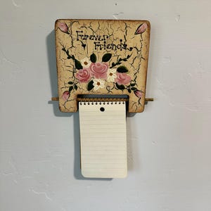 May include: A wooden wall-mounted notepad holder with the words "Forever Friends" and a floral design. The holder has a distressed finish and a small notepad attached with a spiral binding. The flowers are pink and white.